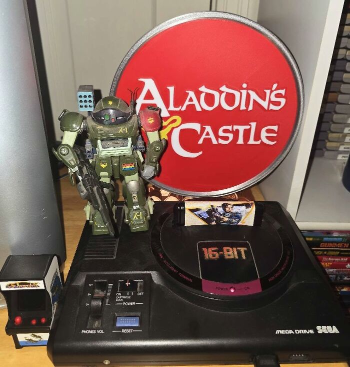 Sega Mega Drive console with 16-bit cartridge, retro gaming robot figure, and Aladdin’s Castle sign in the background.