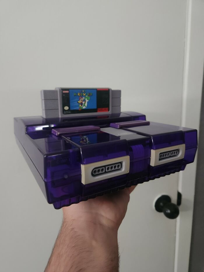 Retro gaming console in purple with a Super Mario World cartridge inserted, held by a hand against a plain background.