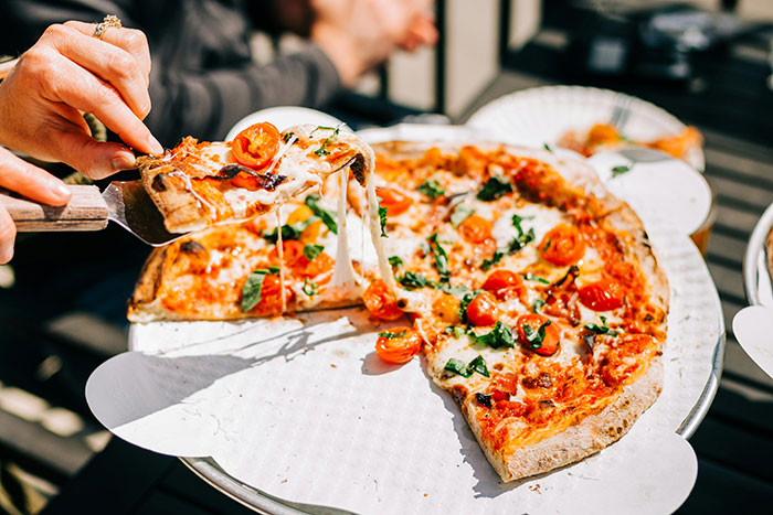 Person serving a gourmet pizza slice topped with cherry tomatoes and fresh herbs illustrating absurd rich people spending.