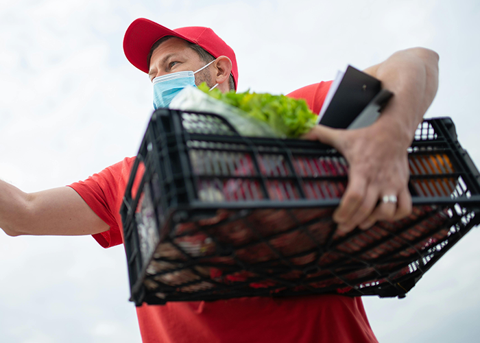 Instacart shopper wearing mask and red cap carrying groceries in a crate, highlighting customer demands and tips.
