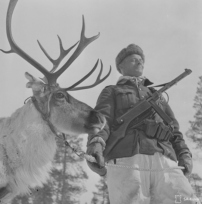 Black and white historical photo of a soldier holding a reindeer by a rope in a snowy forest setting.