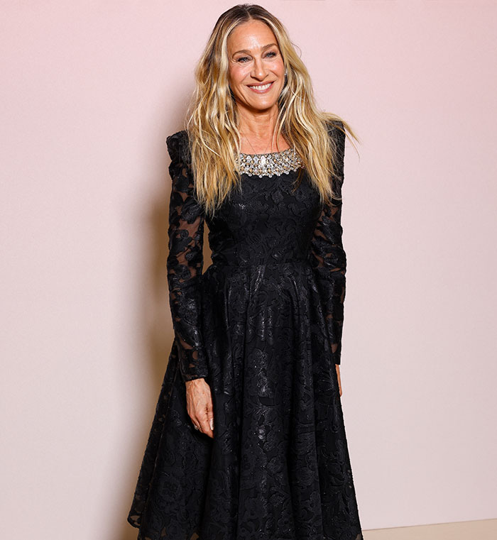 Sarah Jessica Parker wearing a black lace dress smiling against a light pink background at a formal event. Sarah Jessica Parker wearing a black lace dress smiling against a light pink background at a formal event.
