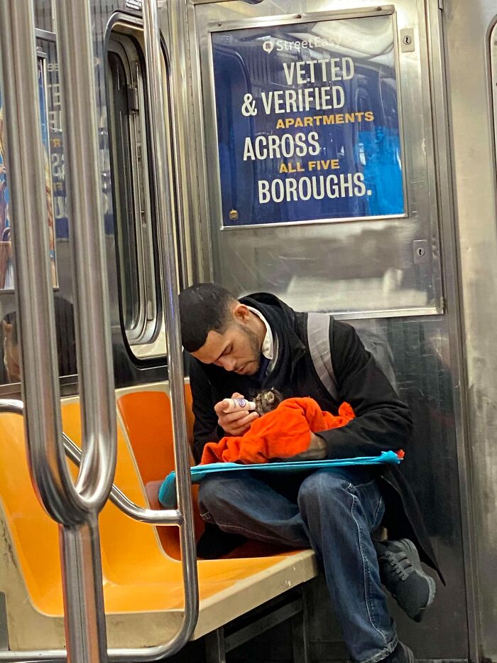 Man feeding kitten wrapped in orange blanket while sitting on subway bench with backpack and jacket.