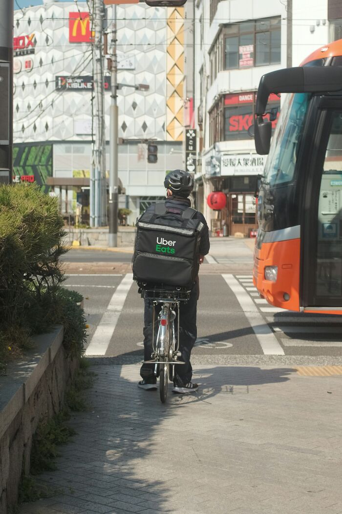 Uber Eats delivery cyclist waiting at a crosswalk in an urban area with nearby bus and commercial buildings scam risks for millennials