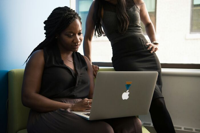 Two women focused on a laptop screen in an office setting, discussing potential scams targeting millennials.