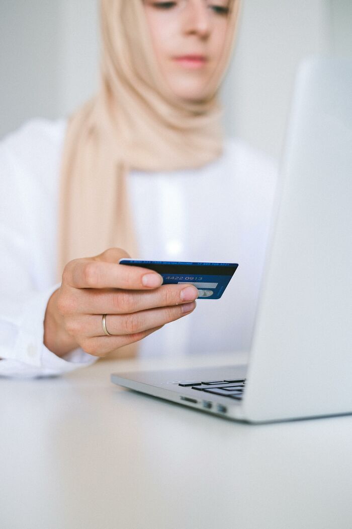 Woman wearing headscarf holding credit card near laptop illustrating scams that fool a millennial online shopping risks.