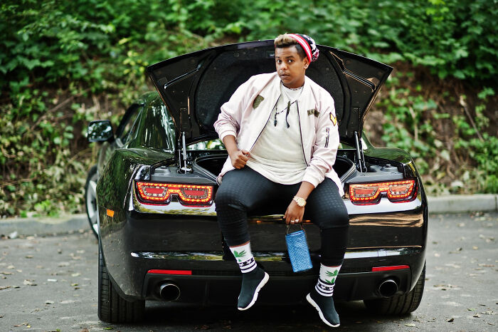 Person displaying alpha vibe sitting on open trunk of a black sports car in an outdoor setting with greenery. - 21