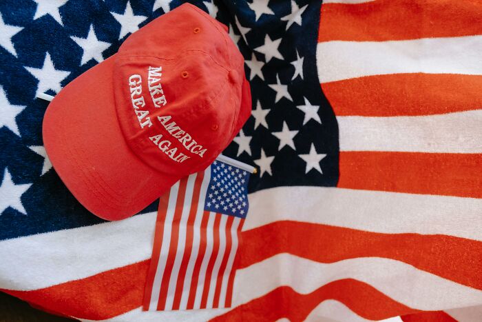 Red Make America Great Again cap and small American flag on a textured American flag background showing alpha vibe traits. - 9