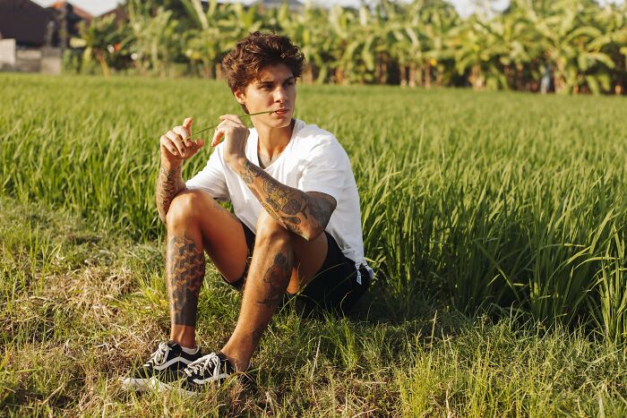 Young man with tattoos sitting in a grassy field, embodying an alpha vibe and confident body language outdoors. - 31