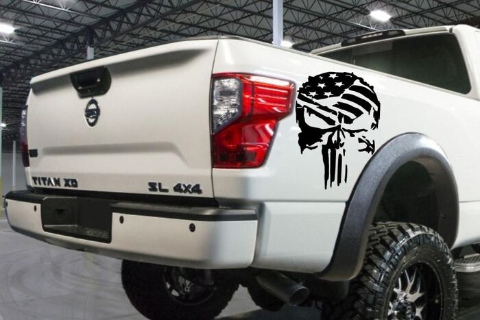 White Nissan Titan truck with Alpha vibe skull decal featuring American flag, parked indoors with raised tires and black fender flares. - 8