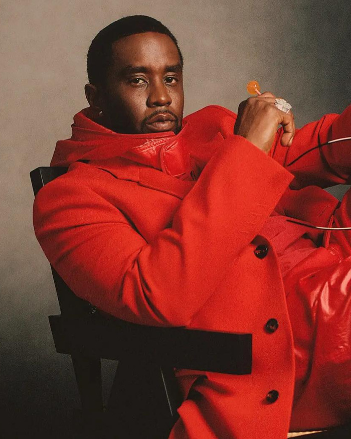 Sean Diddy Combs wearing a red coat, seated on a chair, looking at the camera with a serious expression.