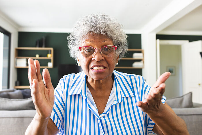 Elderly woman with gray curly hair and glasses explaining wedding registry ideas including a coffin in a modern living room.