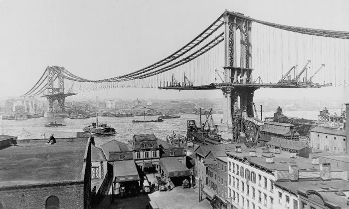 Black and white historical photo showing construction progress of a large suspension bridge over a river.