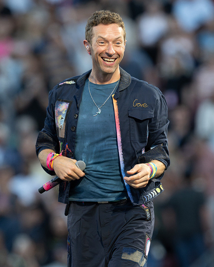 Chris Martin performing onstage, smiling and holding a microphone as fans speculate on Shakira and Chris Martin romance.