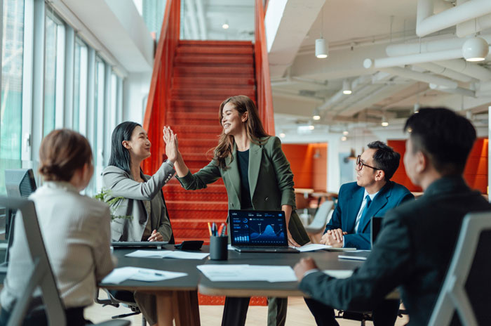 Team members in a modern office sharing inspiration and high-fiving during a meeting with laptop showing data charts.