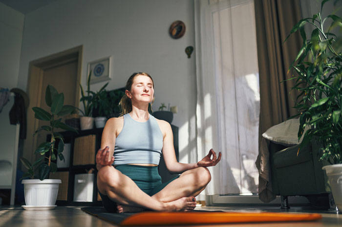 Woman meditating in a sunlit room surrounded by plants, embodying calm and inspiration through mindful practice.
