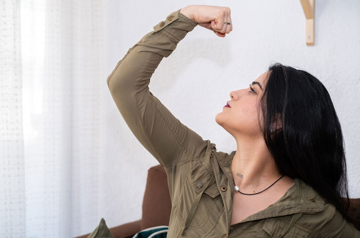 Young woman in an olive jacket flexing her arm indoors, representing motivation and inspiration with short quotes.