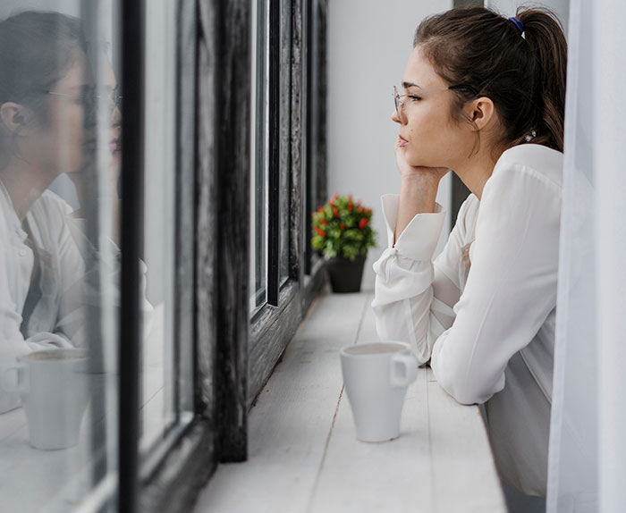 Woman looks thoughtfully out window with coffee cup nearby, reflecting on husband cheating and shocking porch confrontation witnessed by neighbor.