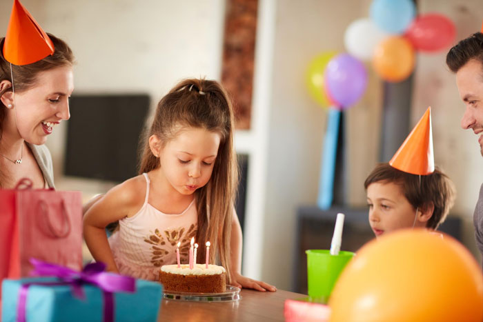 Young girl blowing out candles on birthday cake while family celebrates with party hats and colorful balloons indoors