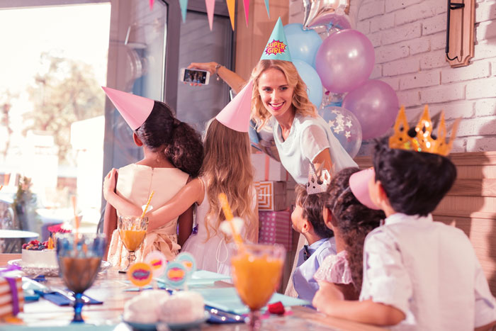 Woman at niece's birthday party taking selfie with children wearing party hats celebrating together indoors.