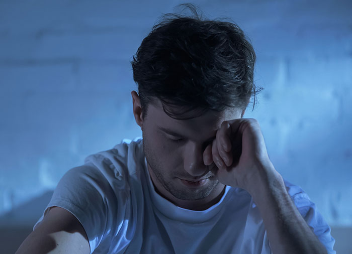 Man in white shirt with eyes closed and hand on face, appearing exhausted and sleepless in a dimly lit room.