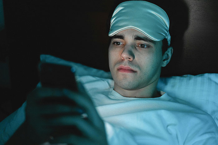 A man lying in bed with a sleep mask on his head, looking worried while using his smartphone at night.