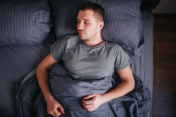 Man lying in bed with eyes closed, appearing to sleep in dimly lit room depicting creepy scene concept.