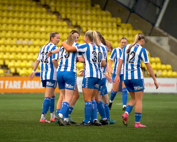 Women&rsquo;s soccer team wearing blue and white striped uniforms huddling on the field during a match in front of empty yellow stands.
