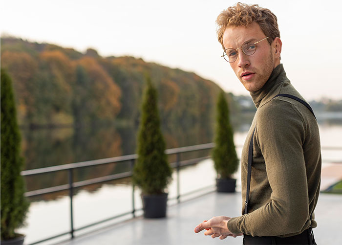 Young man wearing glasses and a turtleneck, quietly observing others near a lakeside terrace with trees in the background.