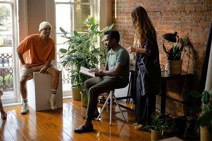 Three people quietly observing and sharing social cheat codes while interacting in a cozy, plant-filled room with exposed brick walls.