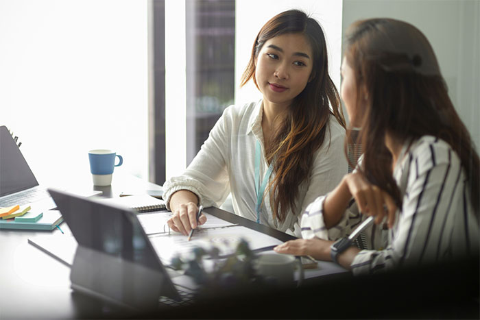 Two women discussing social cheat codes while working collaboratively in a bright office setting with laptops and documents.