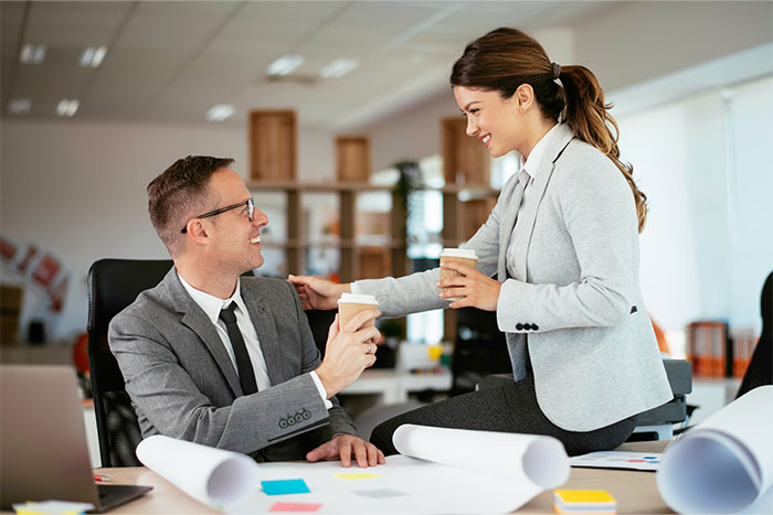 Two coworkers sharing coffee and smiling in an office, demonstrating social cheat codes learned by observing others.