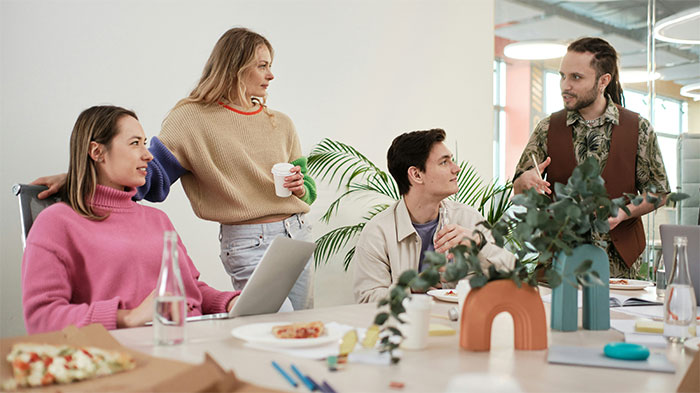 Four coworkers discussing social cheat codes while gathered around a table with laptops, food, and plants in a modern office.
