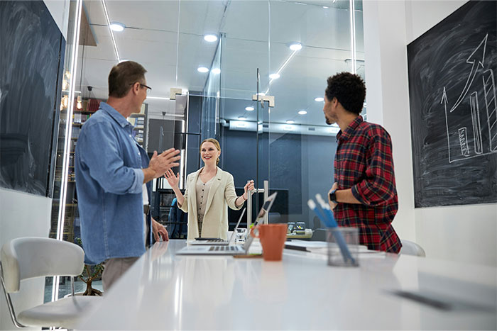 Three colleagues discussing social cheat codes during a meeting in a modern office with charts on blackboards.