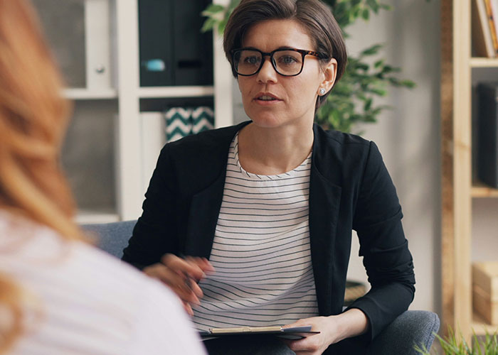 Woman wearing glasses and a black blazer listens attentively during a counseling session about a dad standing by gay son.