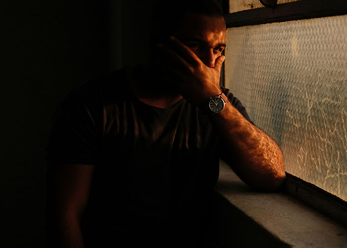 Man sitting alone by window in low light, reflecting on family support after coming out as gay son.