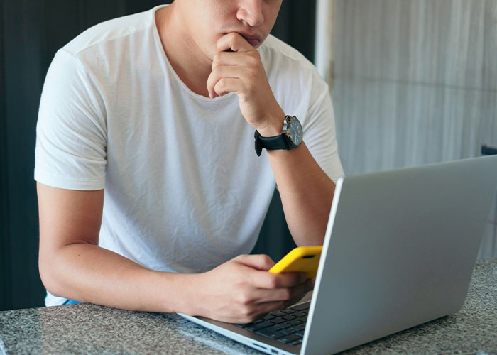 Young man in white shirt holding phone, looking thoughtful while using a laptop, representing dad stands by gay son concept.