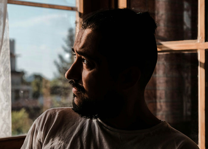 Man with beard looking out window thoughtfully in dim light, representing dad standing by gay son during family conflict.