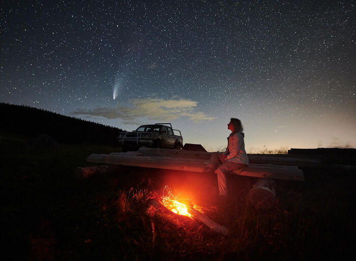 Person sitting by campfire under a starry sky with a comet, illustrating space is a lot scarier than we think.