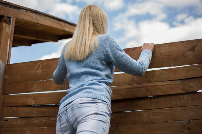Woman in blue sweater peeking over wooden fence, capturing the moment neighbor stealing fruit from garden.
