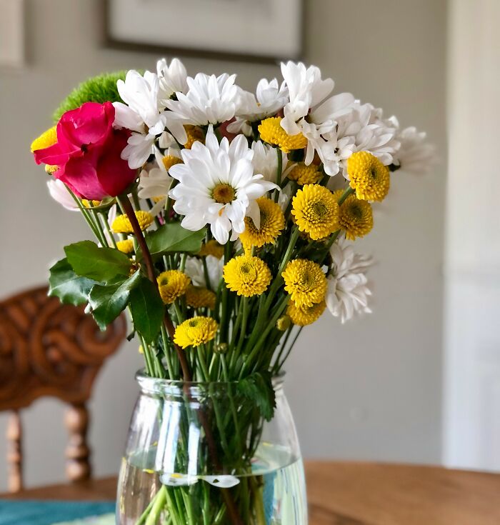 A vibrant bouquet of daisies, yellow chrysanthemums, and a single pink rose in a clear glass vase on a wooden table.