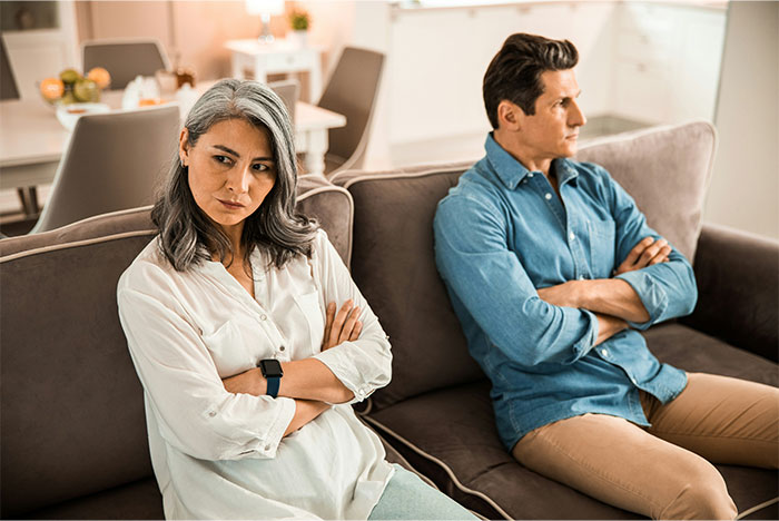 Wife and husband sitting on couch with arms crossed, showing tension and resentment during a serious moment.