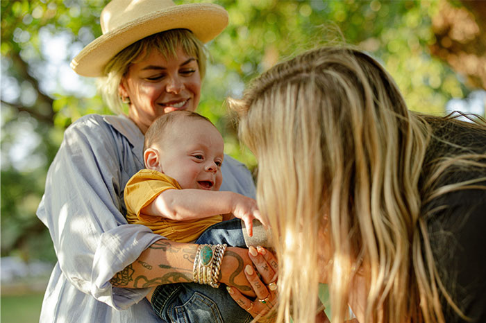 Mother holding baby outdoors while another woman interacts, capturing moments of family and connection on a reluctant trip.