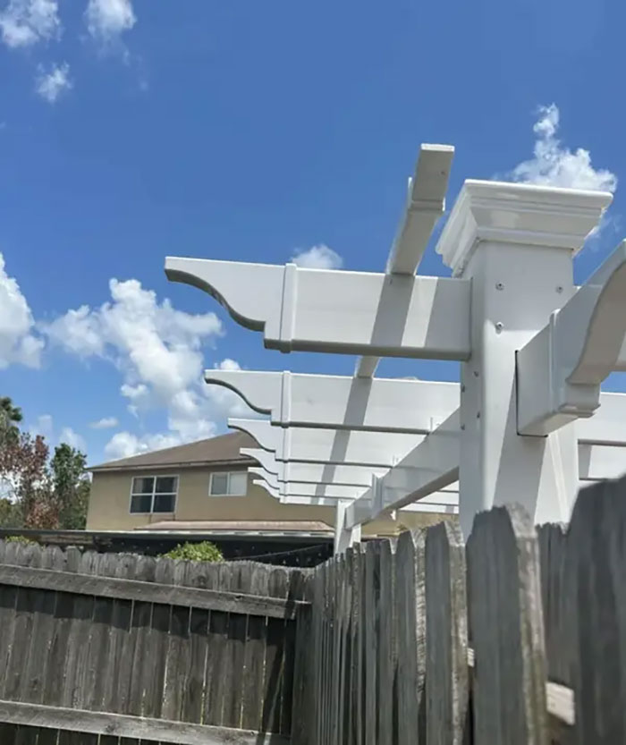 White wooden pergola beams improperly cut over backyard fence under a blue sky, showing selfish people making life harder.