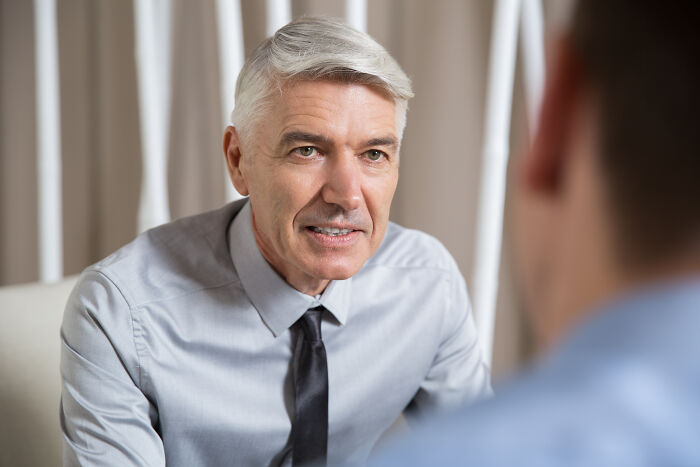Mature hiring manager in a gray shirt and black tie attentively talking to a candidate during a job interview meeting.