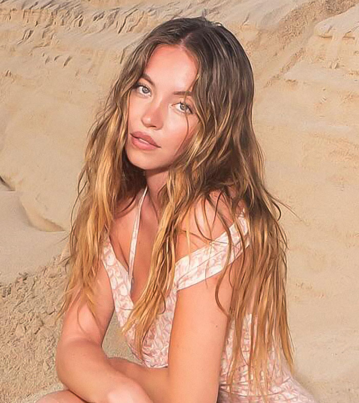 Young woman with long wavy hair in a pink dress posing against a sandy background, reflecting genes with bad taste theme. - 1