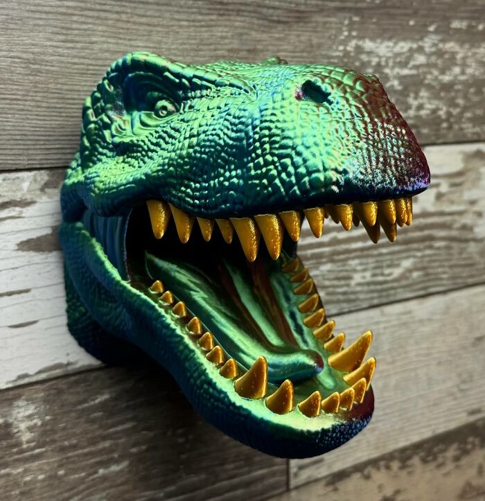 Green dinosaur head wall mount with open mouth and sharp golden teeth displayed on a wooden background.