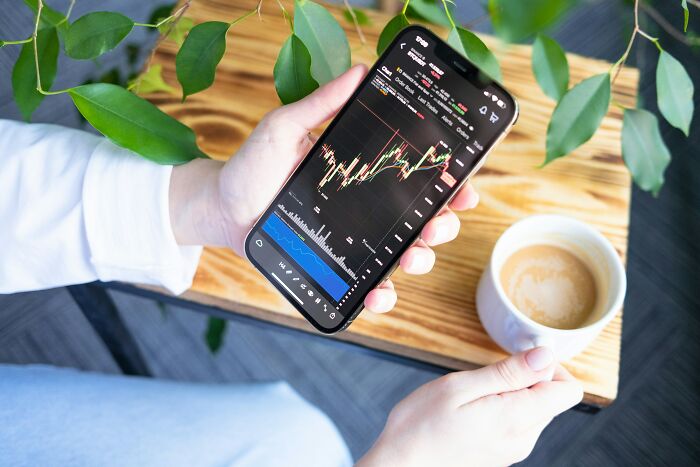 Person holding a smartphone displaying a stock market chart while holding a cup of coffee on a wooden table.