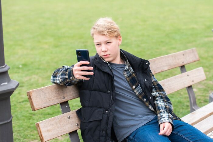 Teen boy sitting on a park bench looking disappointed while checking his phone after a savage rejection story.