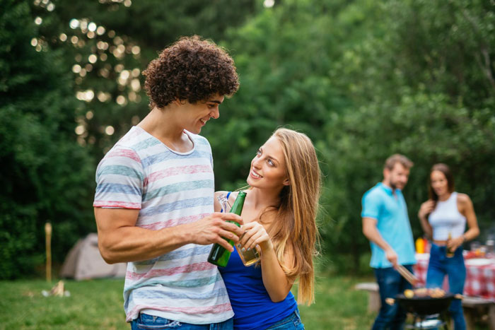 Young couple sharing drinks outdoors, while others barbecue in the background, reflecting on sister's boyfriend seeming quite fake. - 1